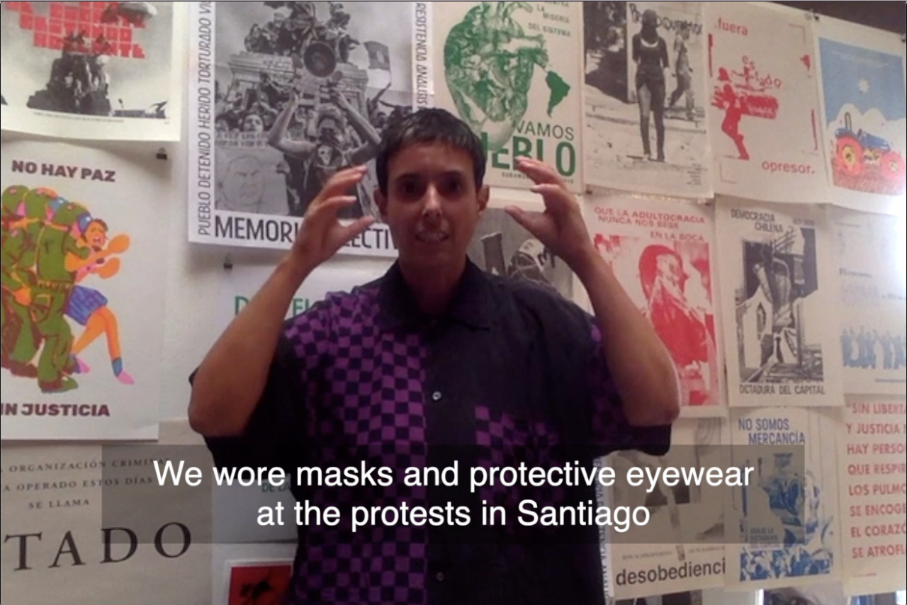 Amelia Bande stands in front of  wall covered with posters and gestures towards her eyes as she says, “We wore masks and protective eyewear at the protests in Santiago.”