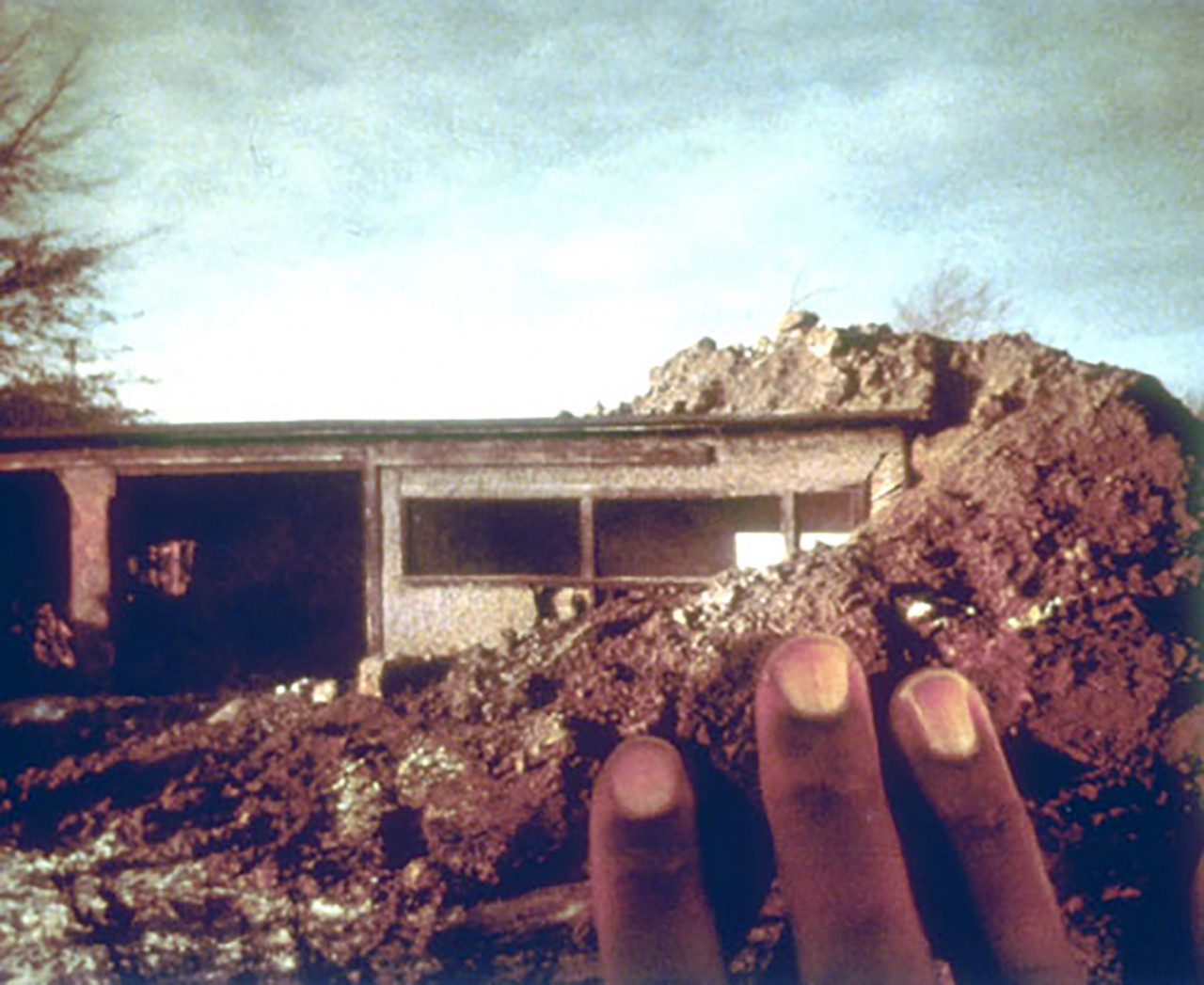 A dilapidated wood shed is shown partially covered by large mounds of earth. Fingers reach out from the lower-right corner to caress the image.