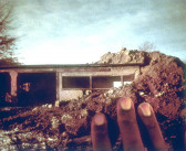 A dilapidated wood shed is shown partially covered by large mounds of earth. Fingers reach out from the lower-right corner to caress the image.