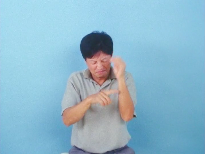 A man wearing a grey polo shirt sitting in front of a sky blue wall signs in Japanese sign language.