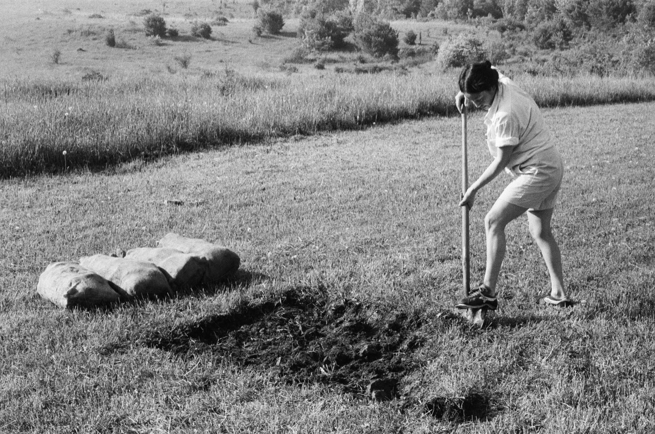 A woman uses a shovel to dig a shallow rectangular pit in a grassy field. Four burlap sacks filled with earth lay to the left side.