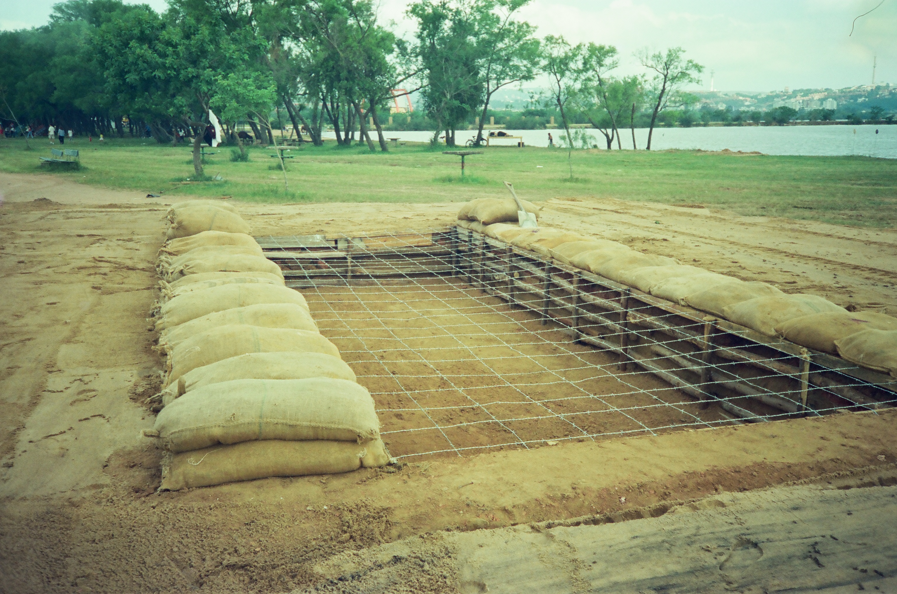 A rectangular pit resembling a mass grave is shown lined with bags of excavated sand. Barbed wire stretches across the pit and a shovel lays to one side. The site of the pit appears to be recreational, with a lake and picnic benches visible in the background.