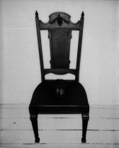 A black and white image of an antique wooden chair with a plush seat. A mold of a human brain emerges from the center of the chair's cushion.