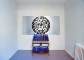 Installation view in a room with white walls and blue floors. In the middle of the wall there is votive candle stand filled with dark blue prayer candles. Above it there is a rectangular artwork with a black and white abstract image in the shape of a circle printed on metal. In the right edge of the image there is a white radiator.