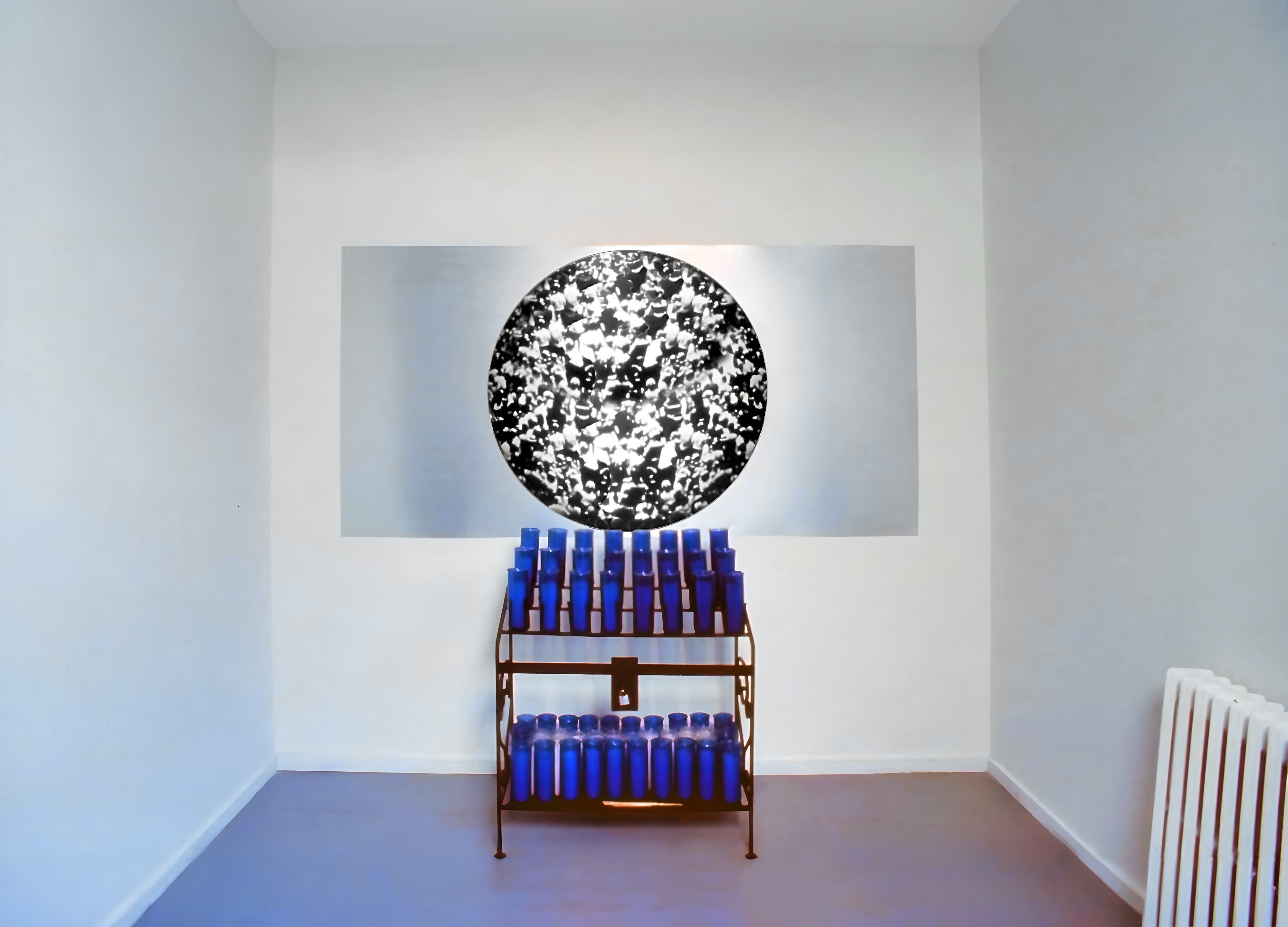 Installation view in a room with white walls and blue floors. In the middle of the wall there is votive candle stand filled with dark blue prayer candles. Above it there is a rectangular artwork with a black and white abstract image in the shape of a circle printed on metal. In the right edge of the image there is a white radiator.