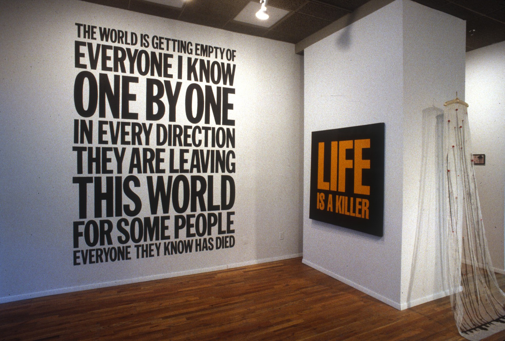 Several paintings and sculptures installed on two walls that form a corner in a gallery space. A square painting featuring large, bold yellow text that reads, 