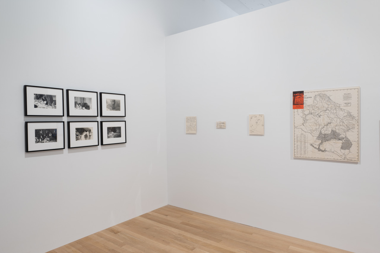 A color image of the corner of a gallery space. On the left white wall, a collection of six framed black and white photographs are arranged in a grid. Each frame is black, with three on the top and three on the bottom. On the right wall are four paper-based items hung in a row. The right-most item is an enlarged map, next to it are three smaller pieces of paper with handwritten notes and drawings.