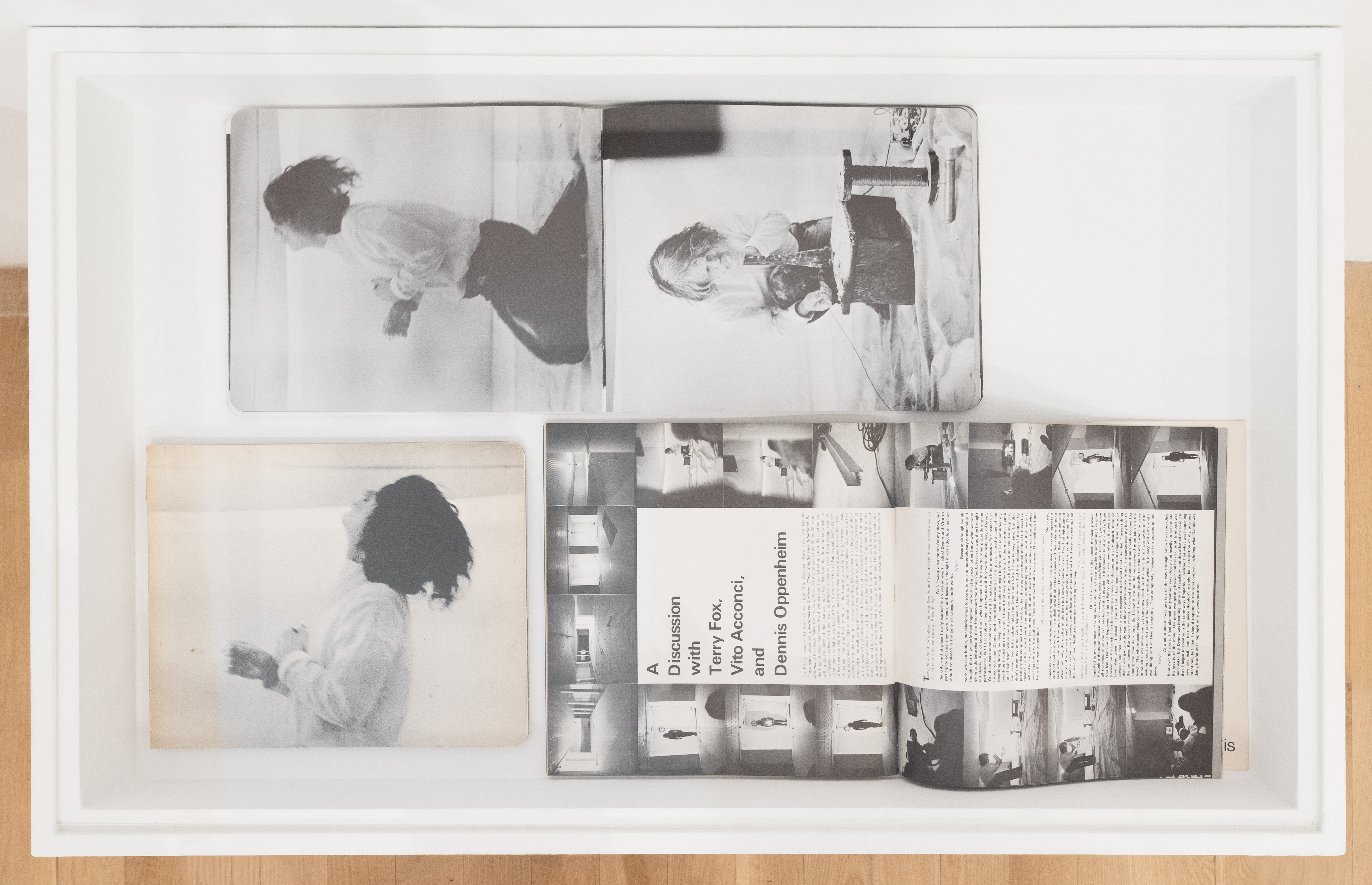  A color image that depicts a bird's eye view of a vitrine. Held within the vitrine is a photobook open to a two-page spread of black and white photographs. The left image is of a man on his knees in a white shirt and black pants with long, dark hair, leaning backwards. The right page is of the same man kneeling in front of a small, low-lying stool. The top of the stool is covered in a white, powdery substance and the man appears to be blowing into a long instrument onto the substance. To the right of the table is a spool of wire. In his free hand, the man holds a string of wire. Below the open photobook is another black and white photograph of the man with dark hair holding his head back and to the left of that image is a two page magazine spread, open to an article titled 