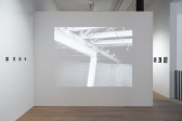 A color image of the corner of a gallery space. On the left wall there is a line of nine polaroid photographs, evenly spaced and hung about five feet from the light wooden floor. The right wall features a frame from a video being played from a projector. The black and white film frame shows the interior of an industrial space, the exposed ceiling rafters and brick wall. 
