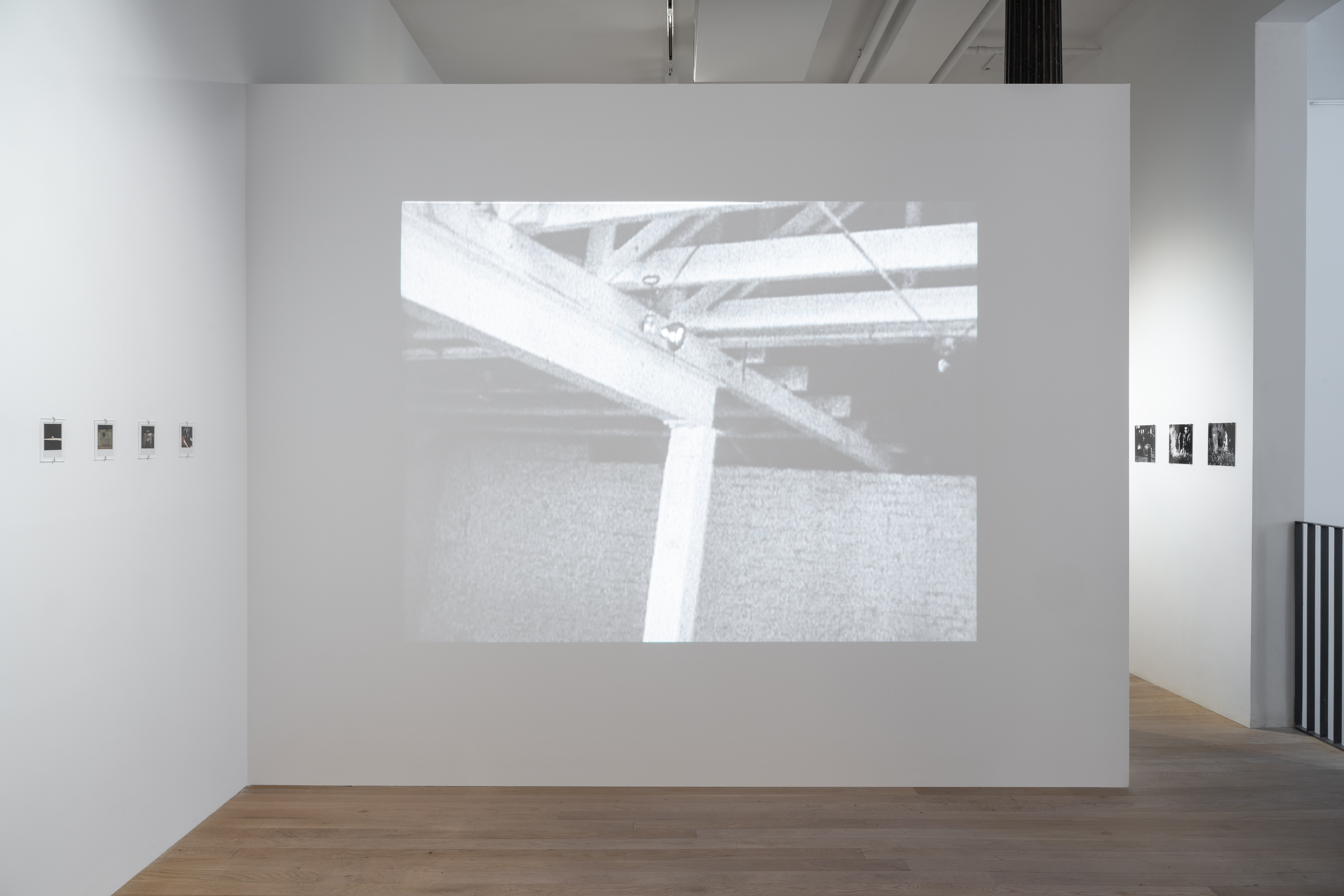 A color image of the corner of a gallery space. On the left wall there is a line of nine polaroid photographs, evenly spaced and hung about five feet from the light wooden floor. The right wall features a frame from a video being played from a projector. The black and white film frame shows the interior of an industrial space, the exposed ceiling rafters and brick wall. 
