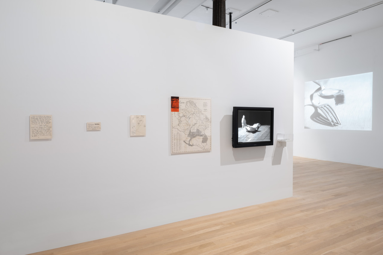 A color image of a freestanding white wall that emerges from the left side of the frame into the gallery space. On the wall, there is a row of six items including two handwritten documents, a drawing, a map, a mounted TV screen displaying a black and white image of a cloth and a small white shelf that holds a plexi box. In the background, a black and white projected image of a fork and spoon is visible on a back wall.