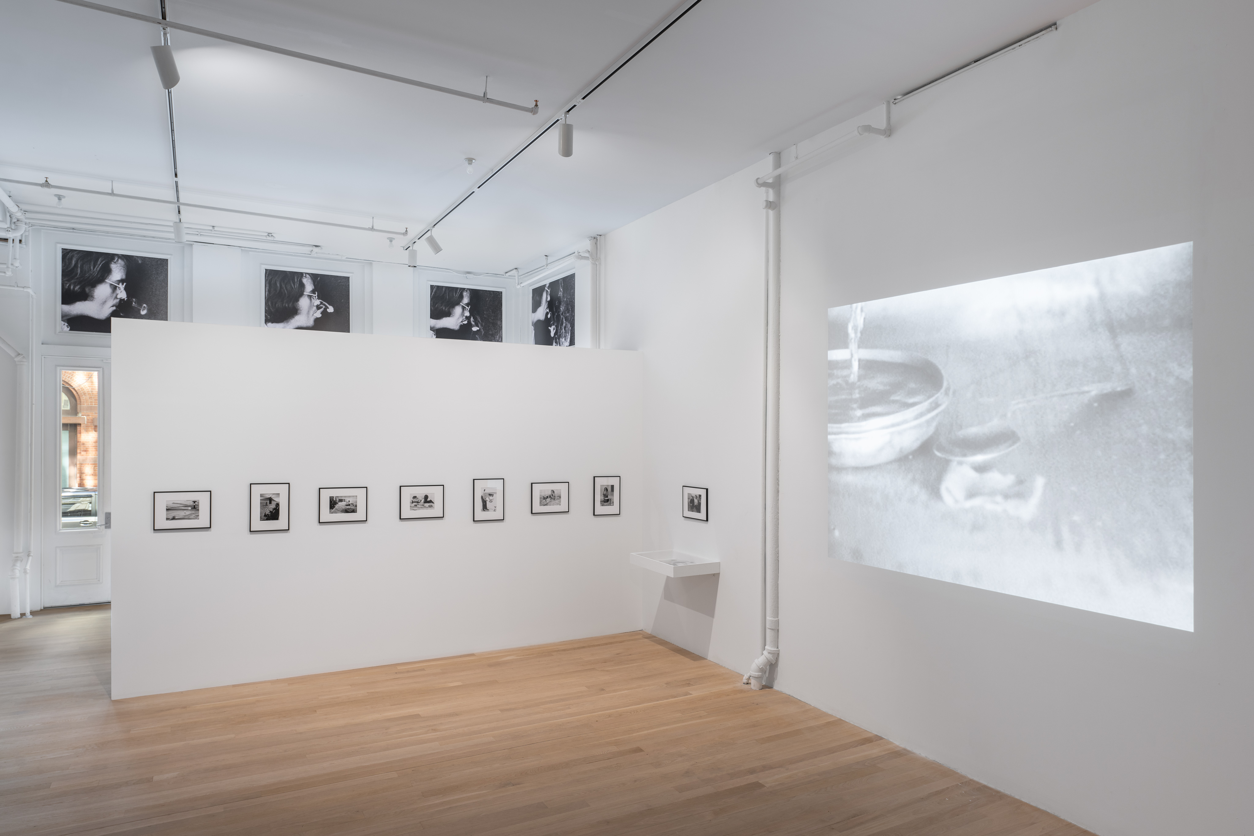 A color image of a gallery space. A freestanding white wall runs perpendicular into an existing white wall. On the right side, projected onto the existing wall, is a black and white image of a spoon resting next to a bowl of water. To the left of the projected image is a single black frame containing a black and white photograph, hung above a small floating vitrine. Installed in a single row on the freestanding wall, is a series of seven black and white photographs housed in black frames. Behind the freestanding walls, a sliver of window that faces the street outside is visible.  Contained within the four transom windows are black and white enlargements of a white man smoking and blowing smoke rings. 