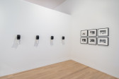  A color image of the corner of a gallery space. A row of four small speakers, evenly spaced and held on small wooden shelves, span the left wall. On the right wall, a collection of six framed black and white photographs are arranged in a grid. Each frame is black, with three on the top and three on the bottom. 