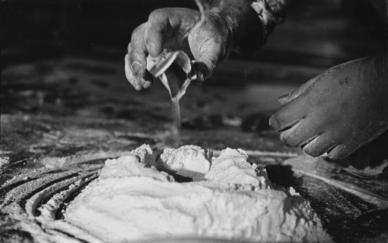 Black-and-white film photograph of a mound of flour on a concrete floor. A figure's hands are shown along the right and top side of the composition. The figure's right hand is poised above the center of the flour mound, pouring yeast into it from a small packet.