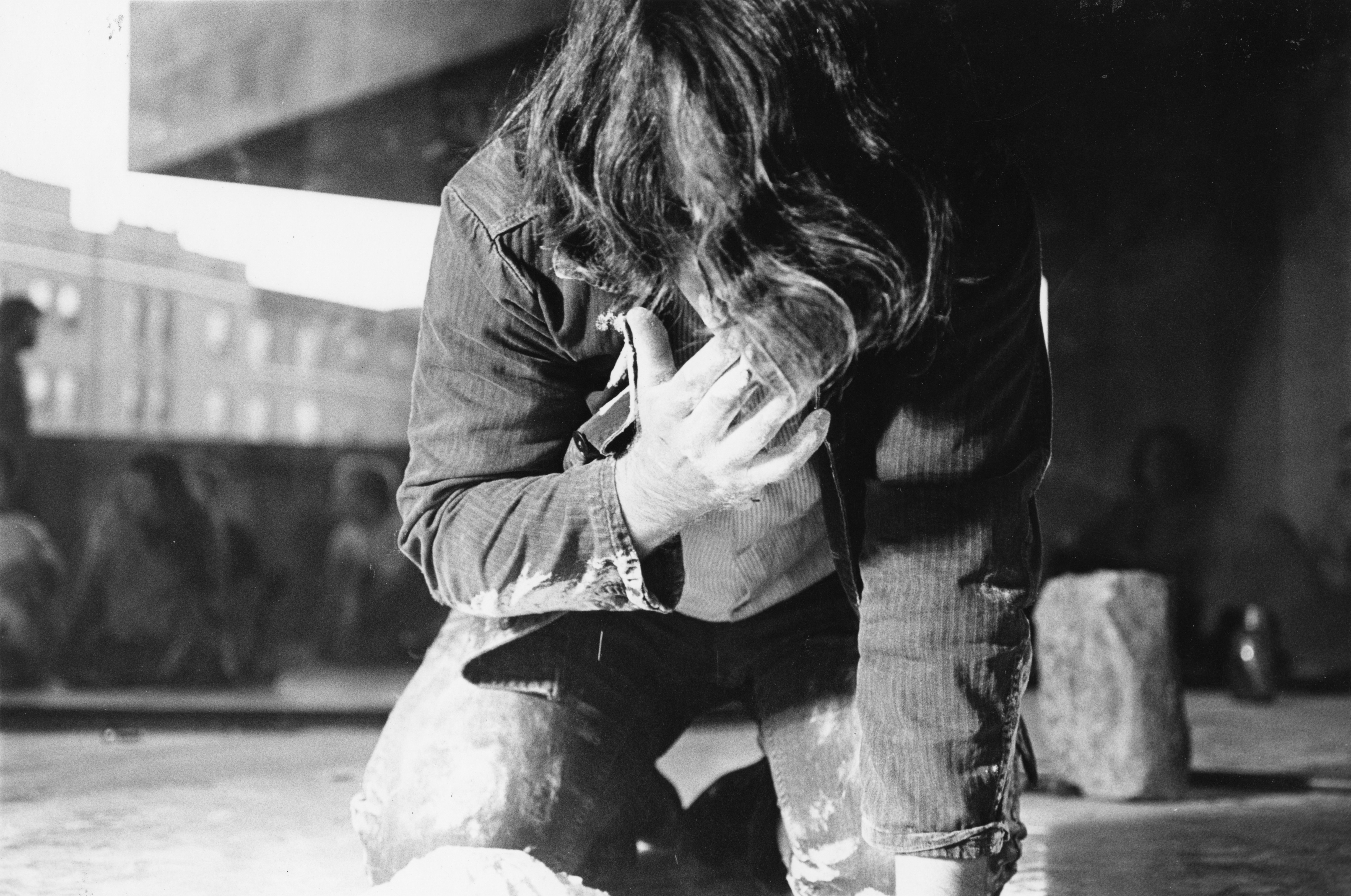 Black-and-white film photograph of a figure bent over, knees on the ground, his left hand reaches up to touch his face which looks downward. His hands and clothing are covered in a dusting of flour. In the background viewers sit on the concrete floor.