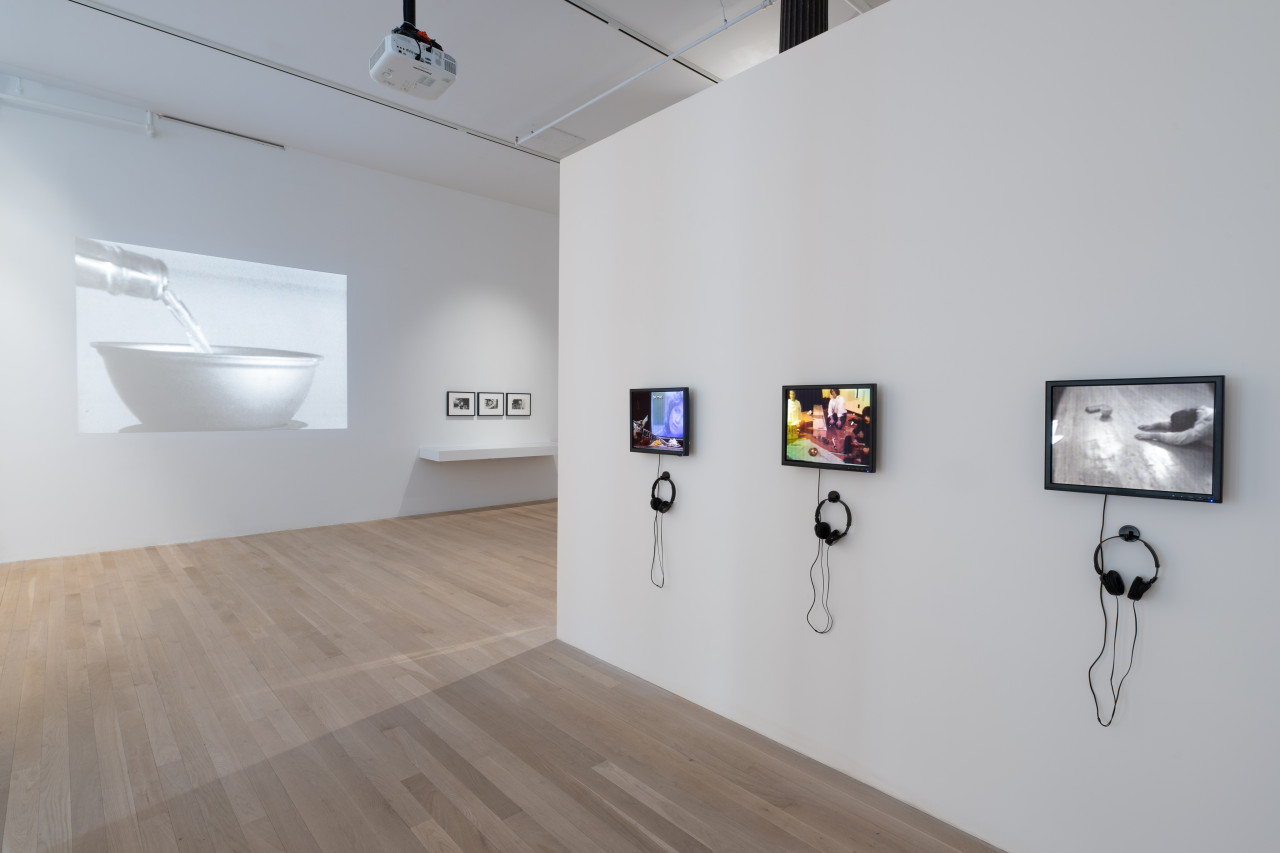 A color image of a free-standing white wall that emerges from the right side of the frame into the gallery space. On the wall, there are three small TV screens, each with a set of black headphones hung on a hook below the screen. The screens on the left and in the middle display color images while the screen on the right displays a black and white image of a man prostrate on the floor. In the background, a black and white image of liquid being poured from a bottle into a bowl is projected onto a white wall. To the right of the projection, three black frames containing black and white photographs are hung above a floating vitrine.