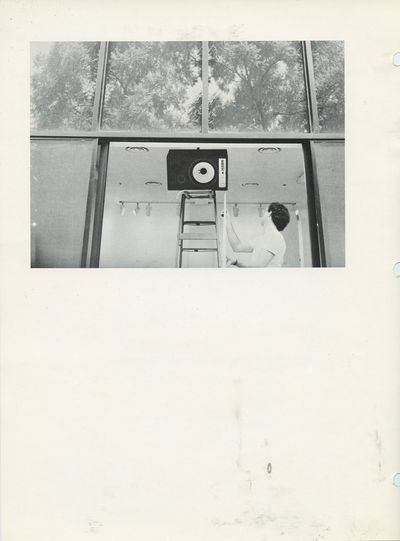 A speaker balances on its side on the top of a step ladder in a gallery space. Directly above the ladder are four large window panes which look out onto trees.