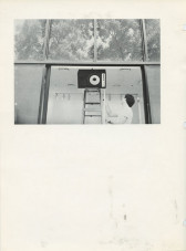 A speaker balances on its side on the top of a step ladder in a gallery space. Directly above the ladder are four large window panes which look out onto trees.
