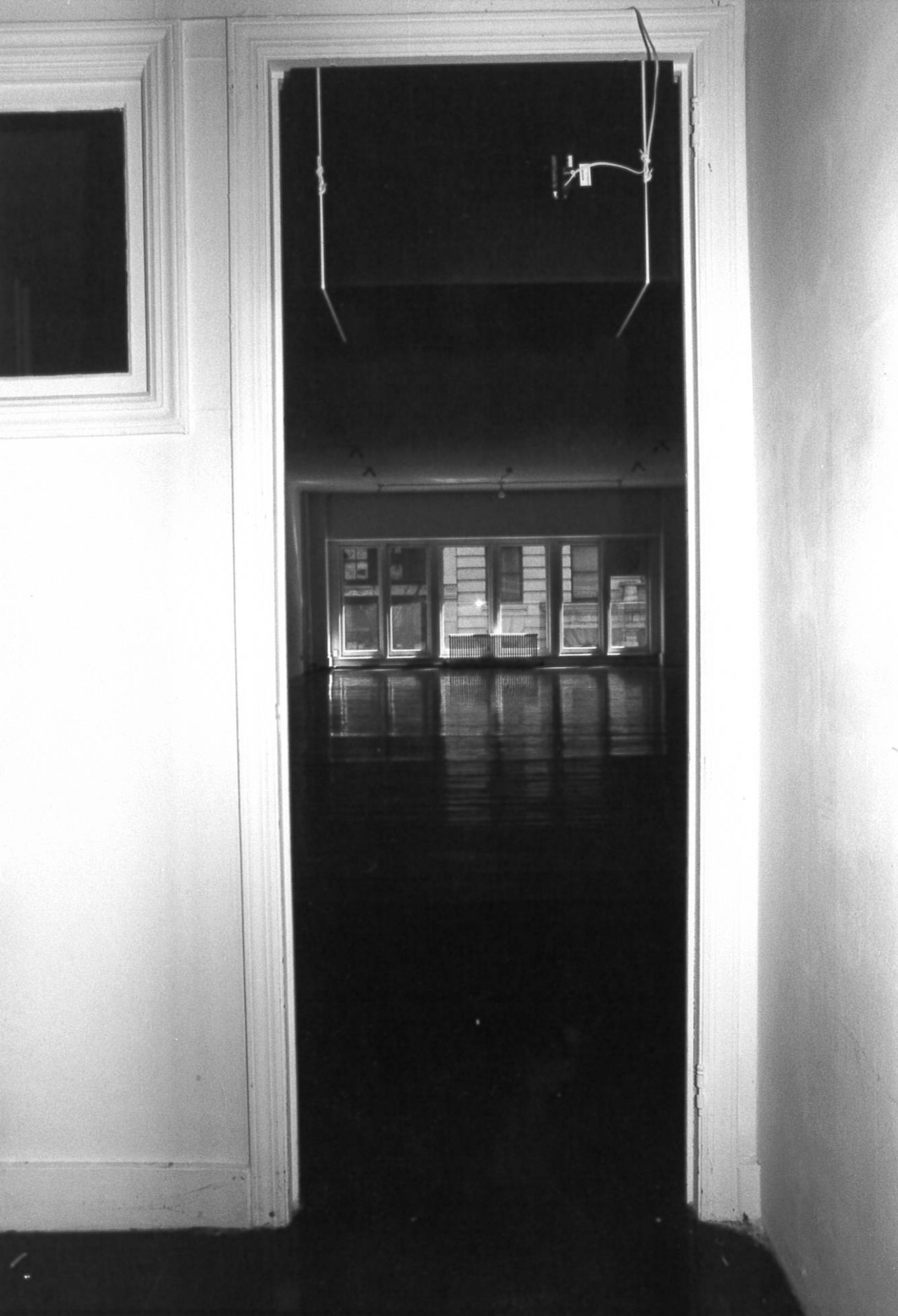 A black and white photograph looking through a doorframe onto several barely illuminated rectangular shapes. A microphone hangs from the top right corner of the doorframe, pointed down towards the floor.