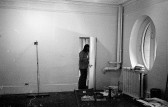 A black and white photograph of a person standing in a doorframe. On the left, a broom rests against the wall. A circular window behind a radiator is visible on the right. Buckets of paint and painting rollers are scattered across the floor.