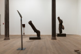 A color image of a gallery space. From left to right: a shovel standing upright on a pedestal, a shovel with an iron claw at the base, a tree log on a pedestal and another tree log on pedestal, with a griffon vulture perched on the branch.