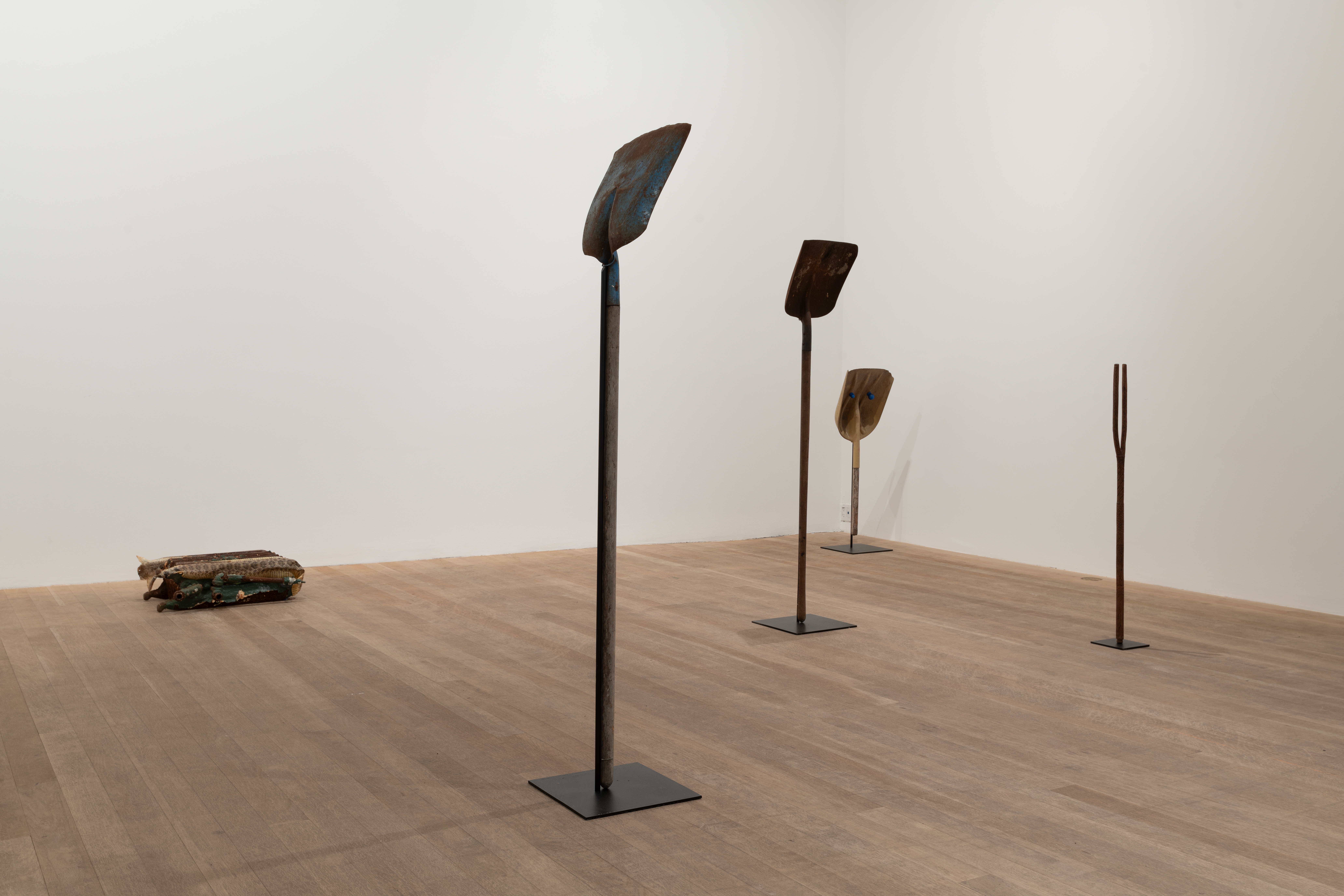 A color image of a gallery space. Three shovels placed upright on pedestals are placed on the floor. To the left, a raditator on its side, partially covered in snake skin, lies on the floor. To the right of the shovels, a wooden divining rod attached to a pedestal sits upright on the floor.