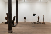 A color image of a gallery space. On the left, two tree log sculptures rest on pedestals. To the right, two shovels standing upright on pedestals flank a taxidermied griffon vulture that sits amongst a pile of concrete rubble. In the center, the top of a shovel afixed to a small childs chair is placed up against the back wall.