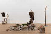 In the center, a taxidermied griffon vulture sits amongst concrete rubble and iron rods spread across the floor in a small pile. To the left, a shovel is afixed to a children's chair and to the right, a shovel is placed upright on a thin pedestal.