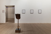 A color image of a gallery space. A sculpture featuring a shovel with a cast iron claw foot sits upright on a pedestal. To the right of the shovel, three framed ink drawings on paper are hung on the wall.