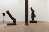 A color image of a gallery space. From left to right: a shovel with an iron claw at the base, a tree log on a pedestal and another tree log on pedestal, with a griffon vulture perched on the branch.