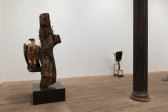 Color image of a gallery space. To the left, a taxidermied griffon vulture rests on a branch protuding from a section of a tree. Rusted, chain link fence is interwoven into the bark of the tree. Off to the right, a small shovel afixed to a childs chair is placed on the ground.