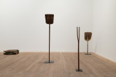 A color image of a gallery space features two shovels, standing upright on pedestals, a radiator sitting on its side; In the forground, a divining rod attached to a pedestal is placed on the floor.