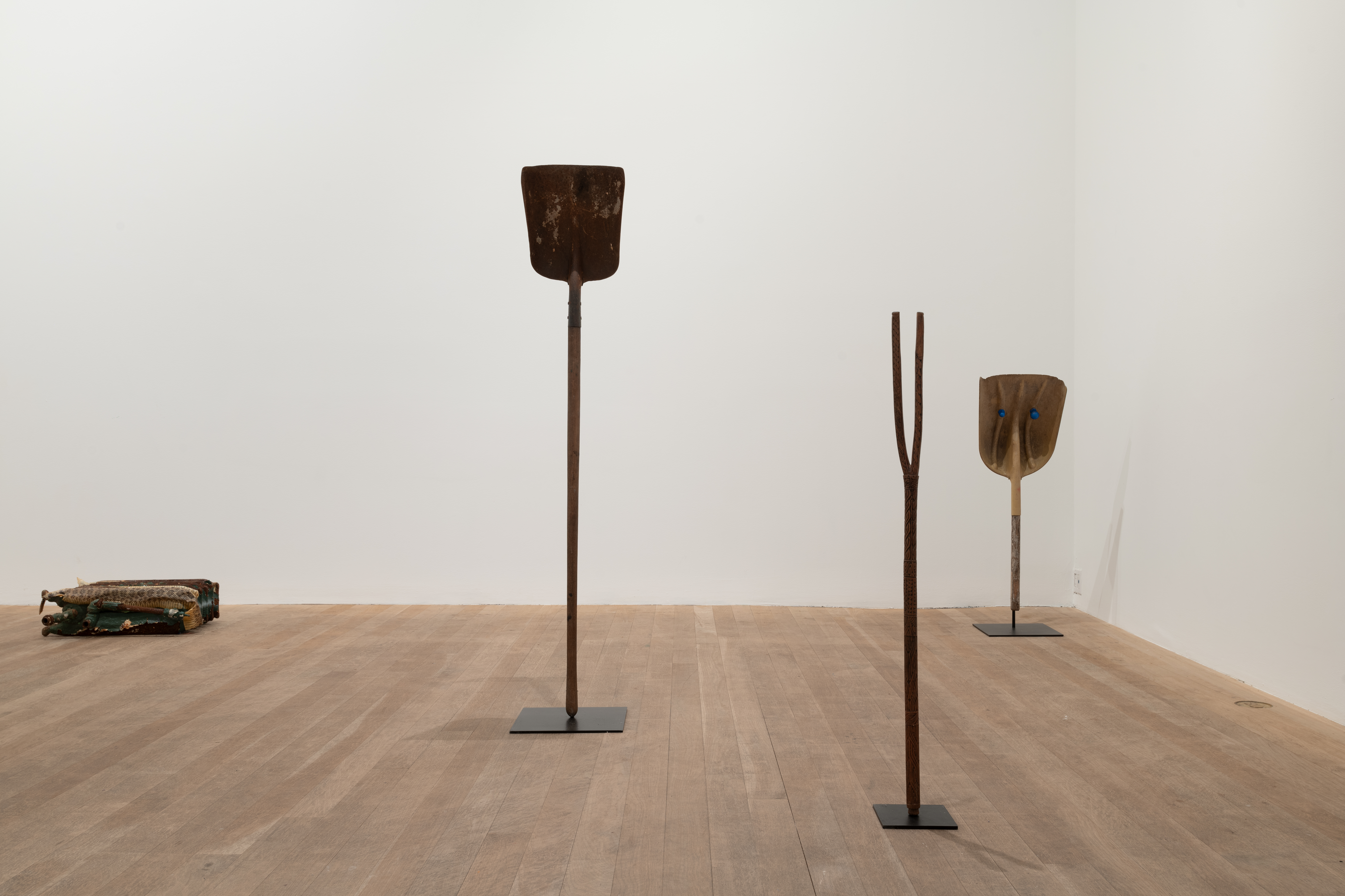 A color image of a gallery space features two shovels, standing upright on pedestals, a radiator sitting on its side; In the forground, a divining rod attached to a pedestal is placed on the floor.