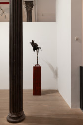 A color image of a gallery space. In the corner of a gallery space, a taxidermied crow is perched atop an iron axe that is decorated with bottle caps. The crow and the iron axe are placed upon a tall pedestal about four feet tall, so that the crow is at, or above the viewers eye level. Entangled in the birds feathers and wings are strands of black hair.
