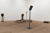 Color image of a gallery space with several sculptures placed on the floor. To the left, a shovel is attached to a child's chair. In the center, a taxidermied griffon vulture is perched upon a pile of concrete rubble that sits in between two shovels standing upright on pedestals. To the far right, a radiator lays flat on the ground.