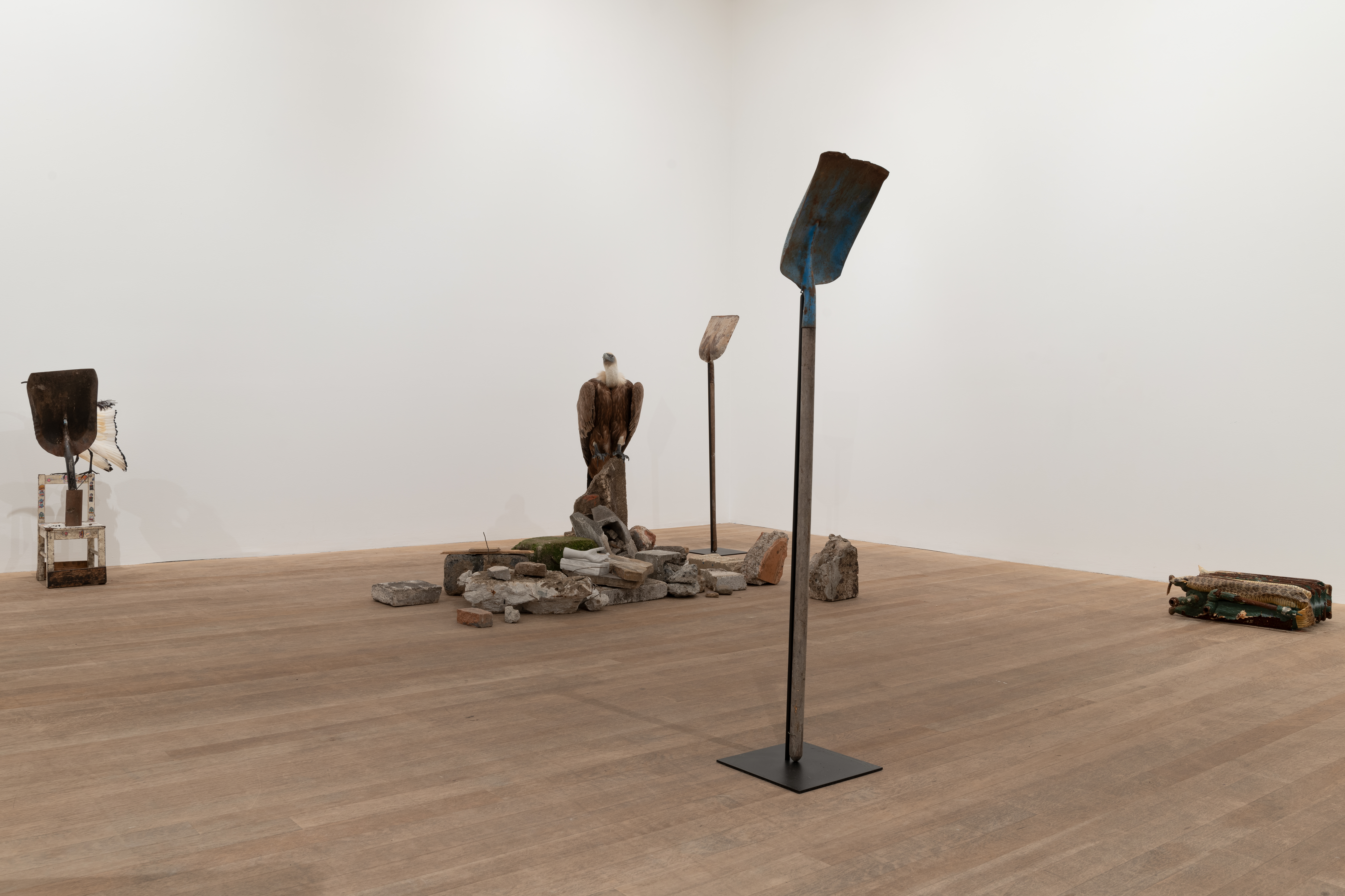 Color image of a gallery space with several sculptures placed on the floor. To the left, a shovel is attached to a child's chair. In the center, a taxidermied griffon vulture is perched upon a pile of concrete rubble that sits in between two shovels standing upright on pedestals. To the far right, a radiator lays flat on the ground.