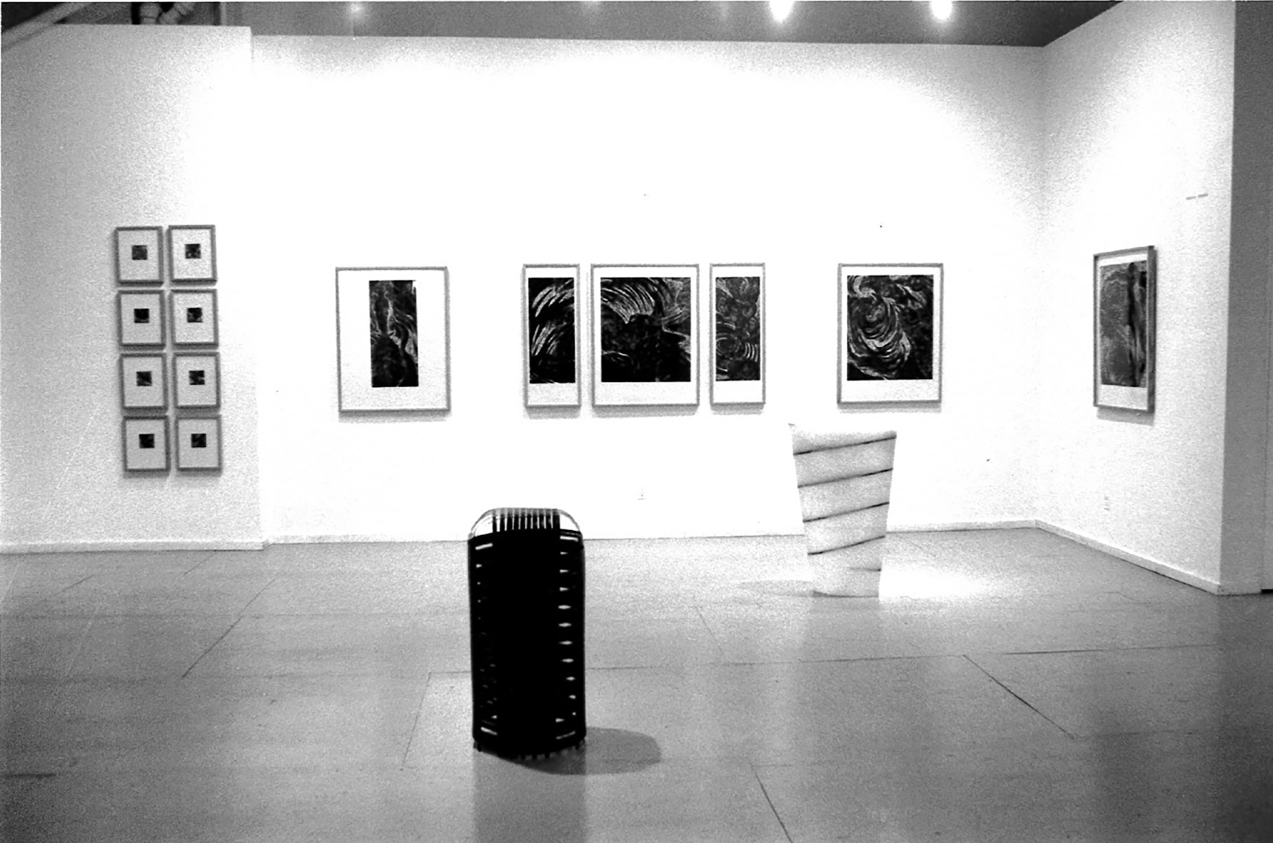 A black and white image of paintings hanging on a white wall, and sculptures occupying the gallery floor.