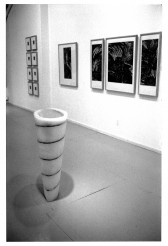 A close up black and white image of multiple paintings hanging on a white wall and a sculpture occupying the gallery floor.