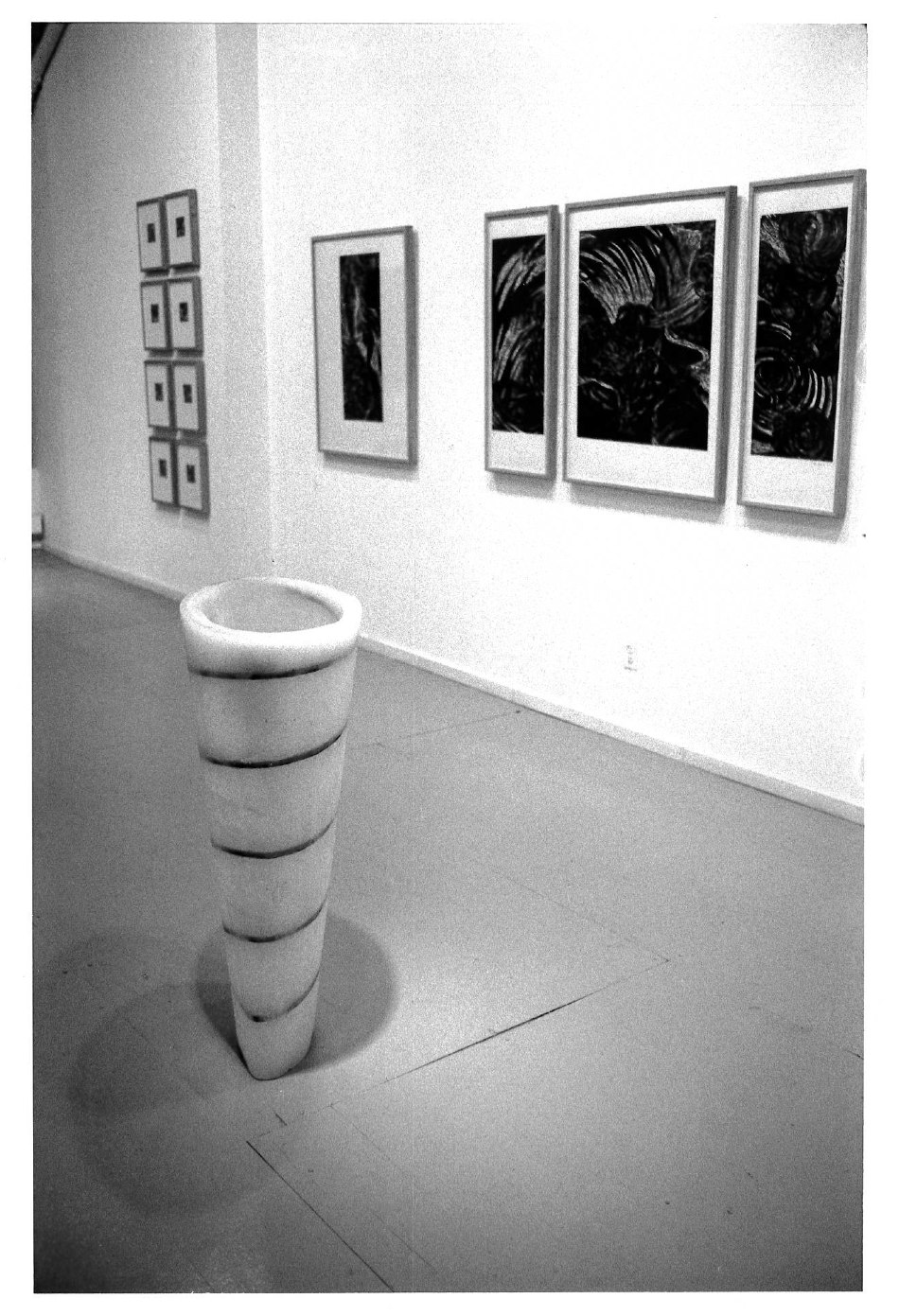 A close up black and white image of multiple paintings hanging on a white wall and a sculpture occupying the gallery floor.