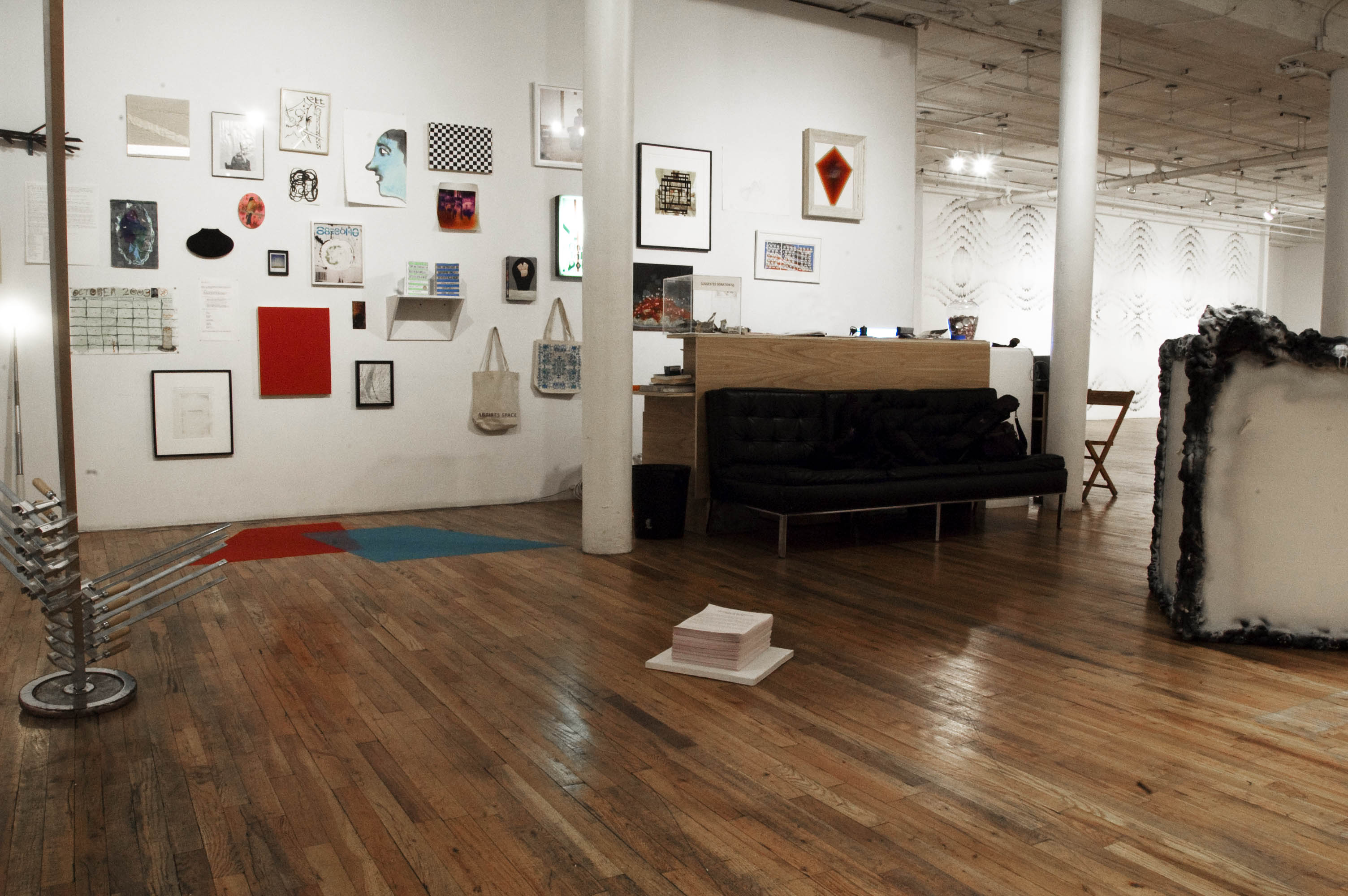 A small stack of papers sits on a white, square tile in a gallery space with wooden floors. Directly behind the stack of papers, a black couch sits back-to-back against a wooden reception desk and counter. Several framed and unframed drawings, photographs, tote bags, and posters hang on the wall behind the reception desk.