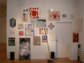 A collection of posters, drawings, paintings, photographs, and collages hang in clusters on a white gallery wall. On the right of the wall is a closed doorway. A long, multicolored poll wedged into a wood stump sits on the gallery floor.
