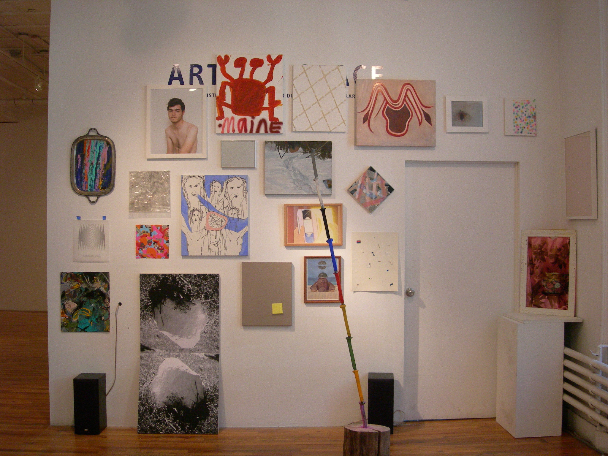 A collection of posters, drawings, paintings, photographs, and collages hang in clusters on a white gallery wall. On the right of the wall is a closed doorway. A long, multicolored poll wedged into a wood stump sits on the gallery floor.