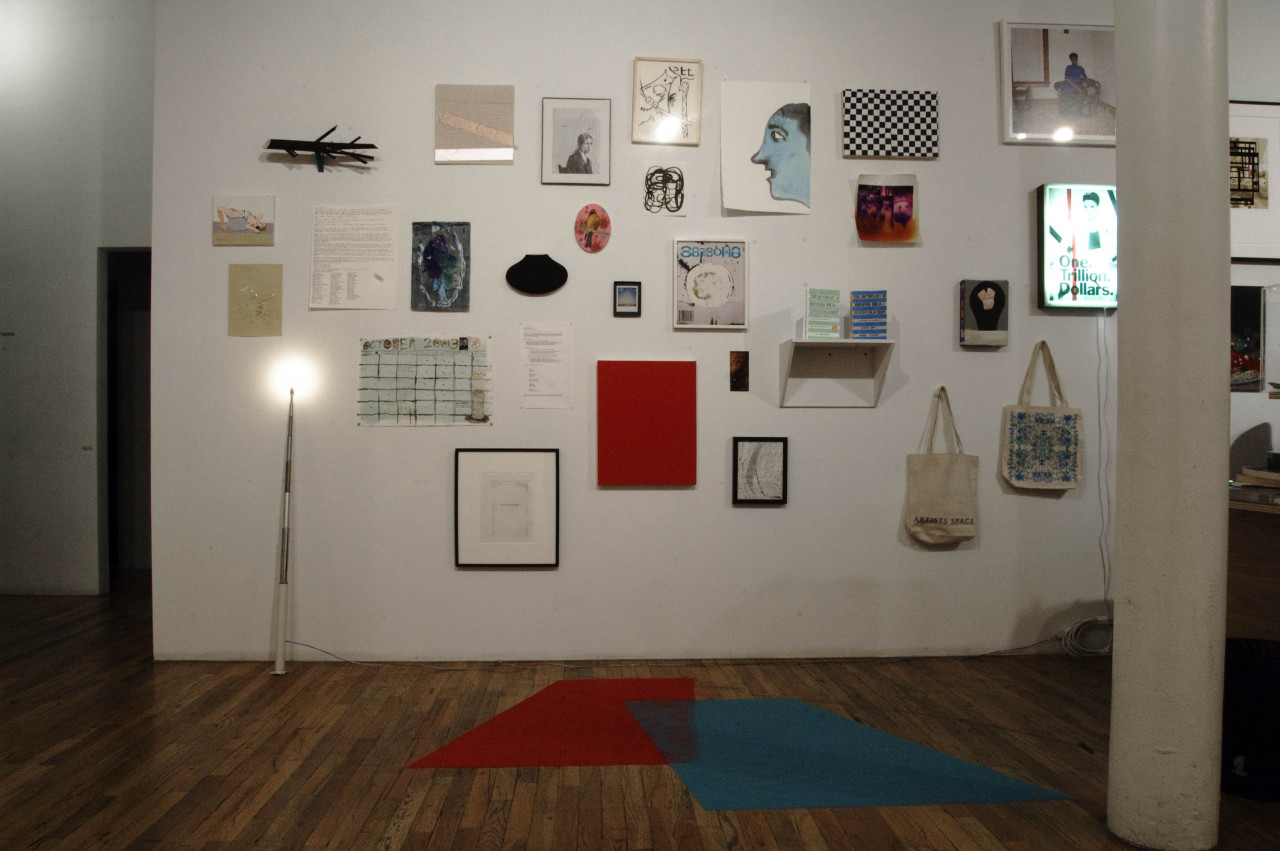 A collection of drawings, paintings, tote bags, and posters hang on a gallery wall. Two geometric, carpet-like shapes are spread across the floor before the wall.