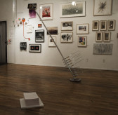 A sculpture consisting of a large, leaning metal pole installed in a gallery space in front of a wall of framed photographs, drawings, and other two-dimensional works. The pole is braced at the floor and leans to the left. Several metal clamps are attached to the bottom portion of the pole, near where it meets the floor. A stack of white papers is displayed on the ground in front of the sculpture.