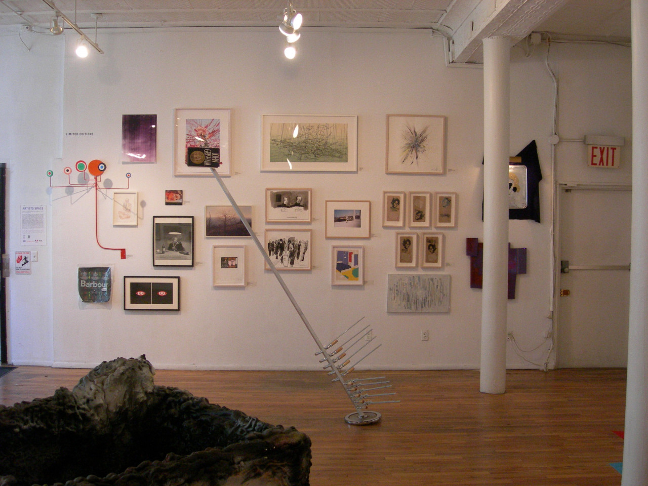 A long, thin metal pole attached to a round base sticks into the air at a jaunty angle in a gallery space. Several metal clamps are attached to the bottom third of the pole. A collection of photographs, drawings, and paintings are framed and hung on the gallery wall behind the metal pole.