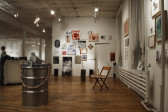 A ceramic figure of a headless gray dog sits on the top of a shining metal barrel in an open gallery space. A garland of sausages connected to the ceramic dog drapes over the sides of the barrel. A wooden chair sits behind and to the right of the barrel. Many paintings, drawings, photographs, and posters hang on the gallery walls.