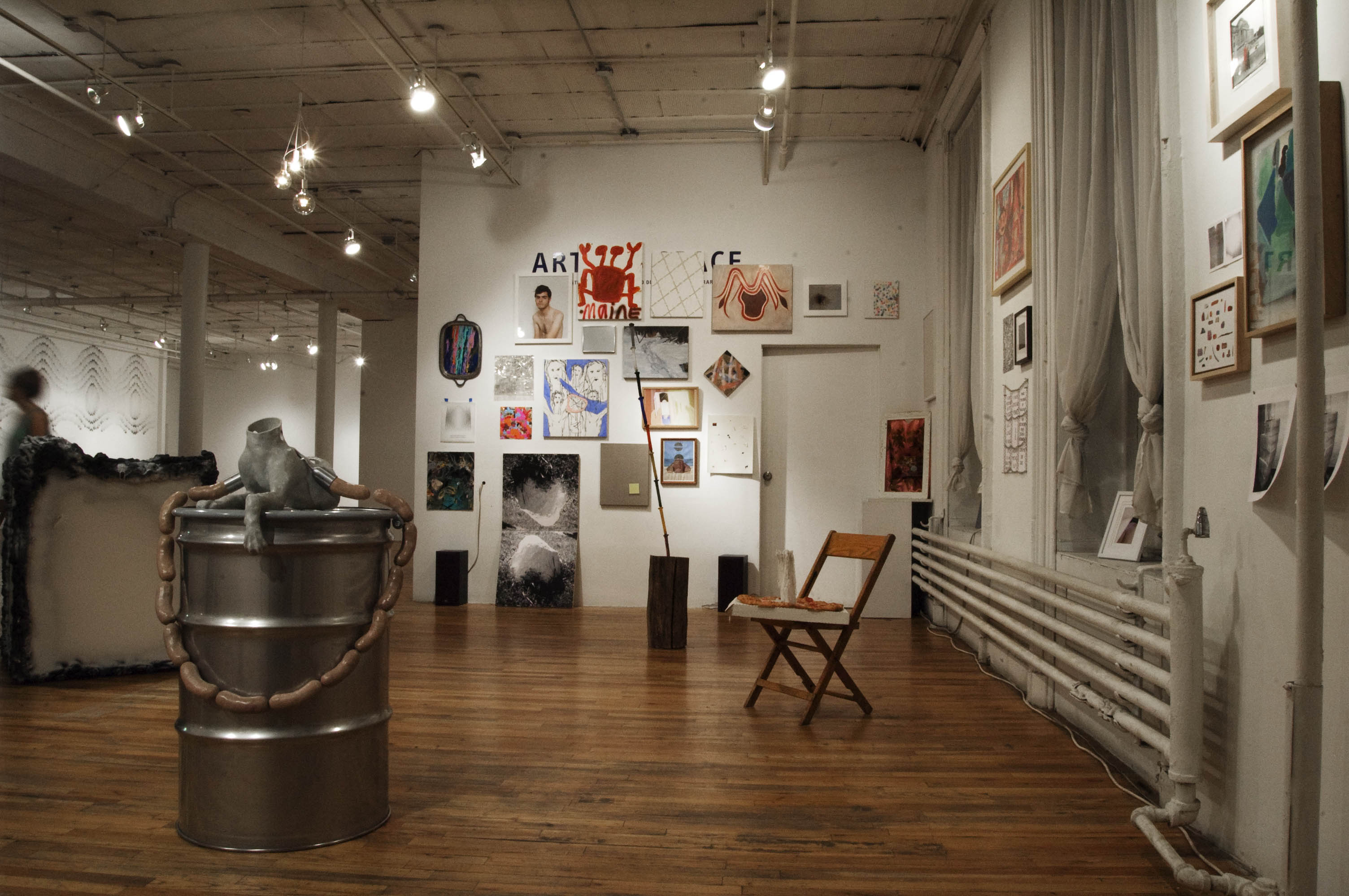 A ceramic figure of a headless gray dog sits on the top of a shining metal barrel in an open gallery space. A garland of sausages connected to the ceramic dog drapes over the sides of the barrel. A wooden chair sits behind and to the right of the barrel. Many paintings, drawings, photographs, and posters hang on the gallery walls.