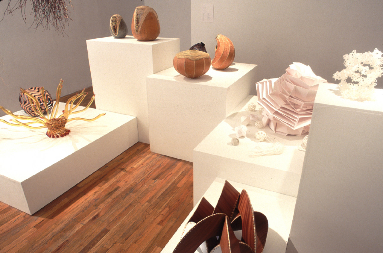 A series of amorphous, basket-like objects sit on a series of plinths of varying heights in a gallery space. The baskets are composed of a variety of materials, including wood, grass, gourd, and paper.