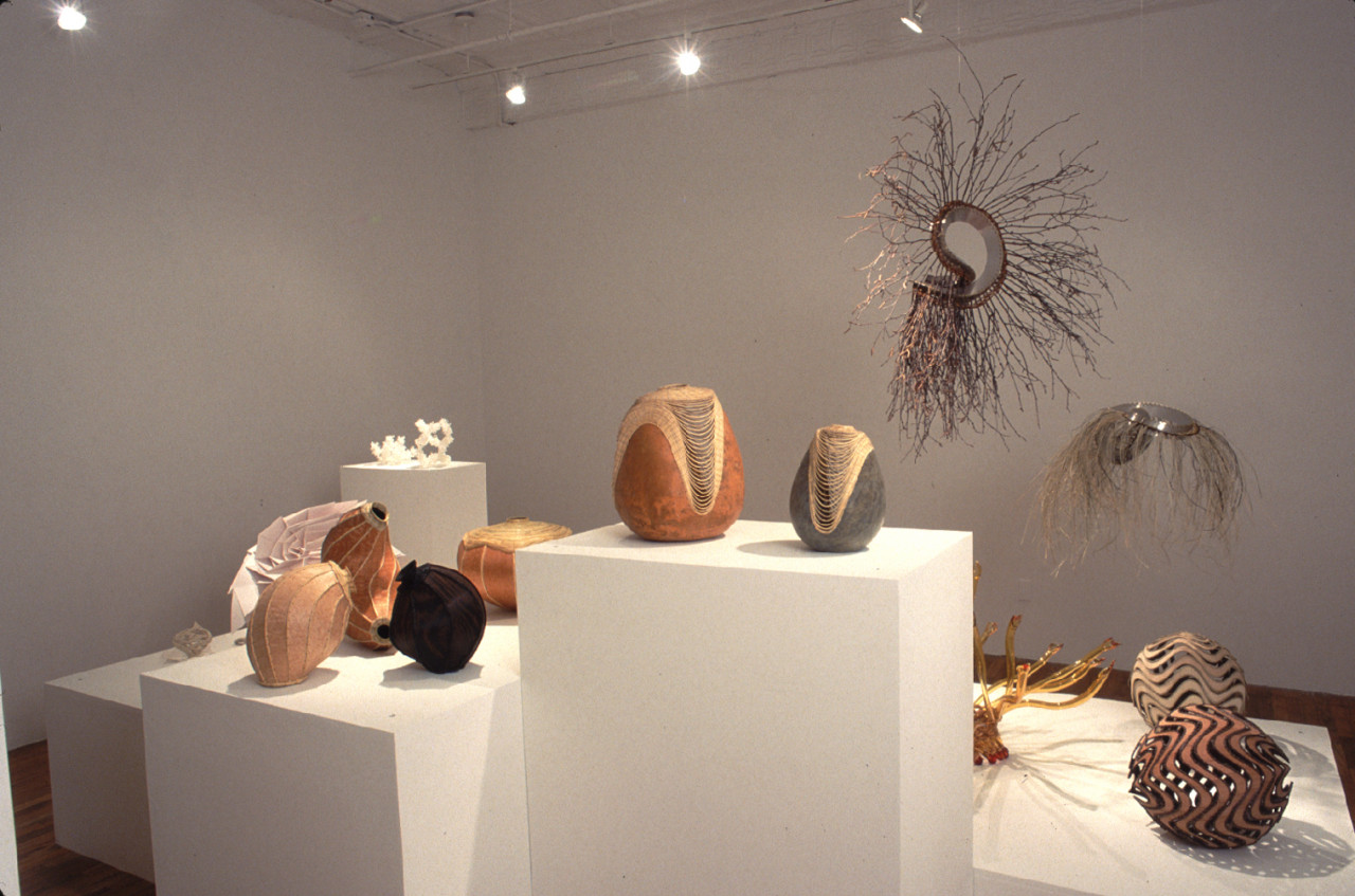 A series of amorphous, basket-like objects sit on a series of plinths of varying heights in a gallery space. Two additional scultural forms composed of sticks and grasses, as well as metallic materials,  hang from the ceiling behind the plinths.