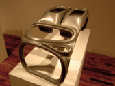 An abstract sculpture with a chrome finish is displayed on a white pedestal. The sculpture's form includes amorphous voids alongside somewhat rectangular structures.
